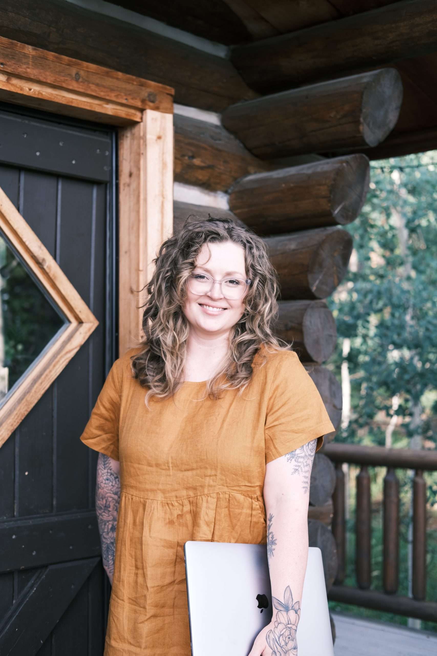How I Became a Web Designer (And Built a Career I Love) | Westco Web Design Colorado web designer in a yellow dress, standing in front of a log cabin, holding a laptop with tattoos on her arns.